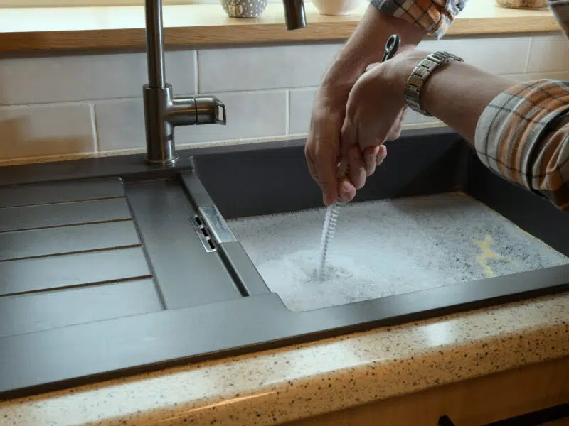A person in a plaid shirt cleans a sink with a brush, surrounded by soapsuds. The kitchen setting has a polished, organized feel.