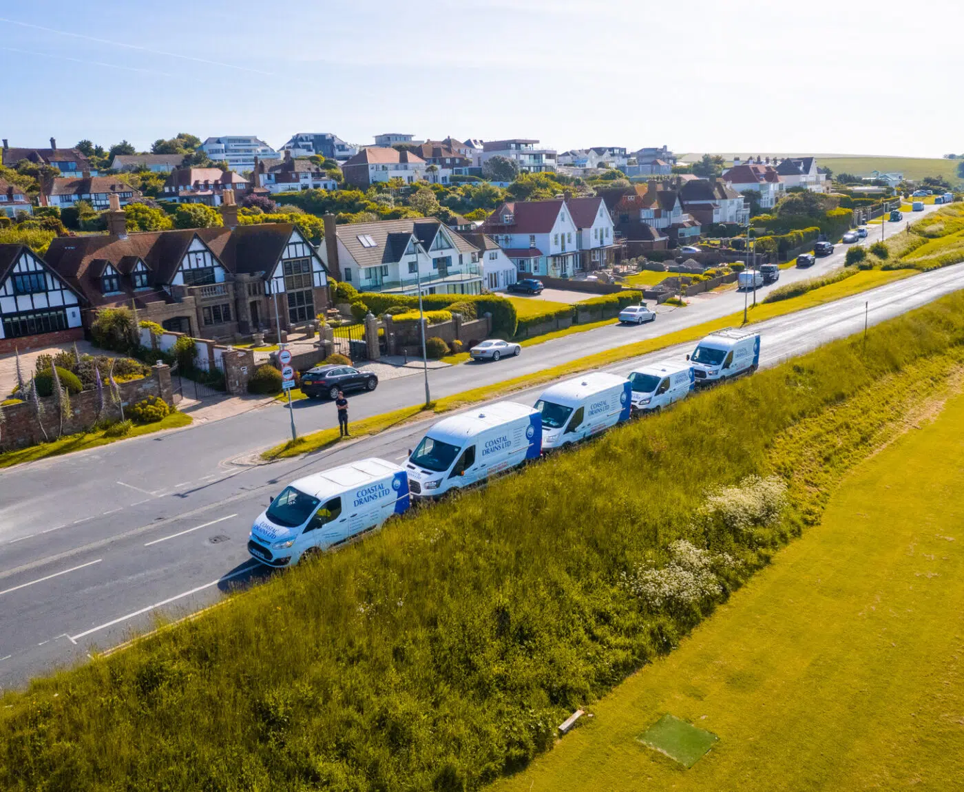 Aerial view of a row of white service vans driving along a coastal road with grass on one side and a row of charming suburban houses on the other.