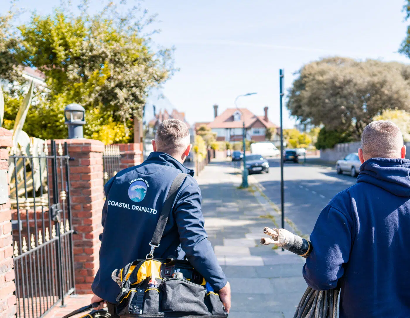 Two workers in blue uniforms walk along a sunny suburban street. One carries tools, the other holds a large hose. Trees and brick walls line the road.