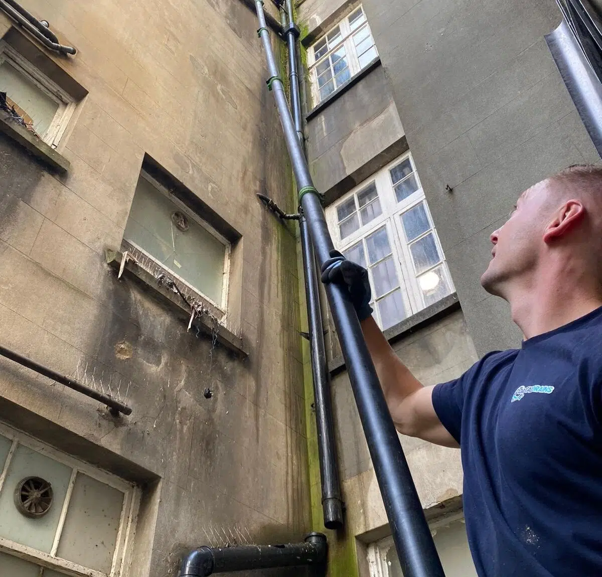 A worker in a navy shirt and gloves uses a long tool to clean a vertical pipe on a tall, weathered building. The wall shows green moss and dirt buildup.