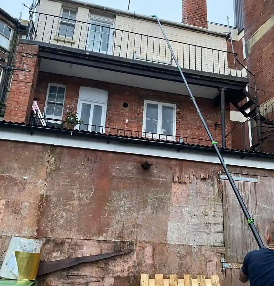 A person using a long pole to clean third-floor windows of an old red brick building. The scene includes weathered walls, wooden pallets, and a green trash bin, conveying a gritty urban atmosphere.